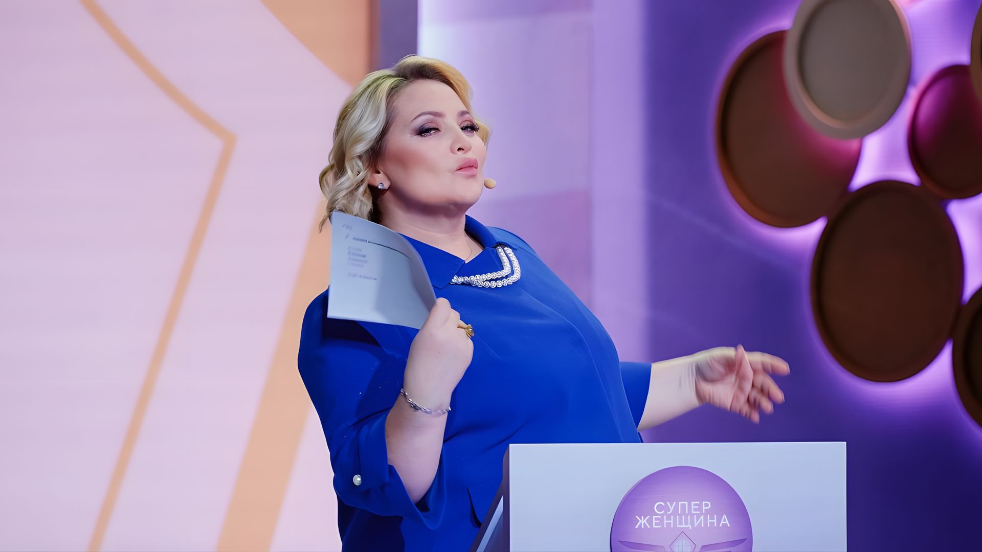 Светлана Пермякова в синем костюме за трибуной обмахивает себя бумагами