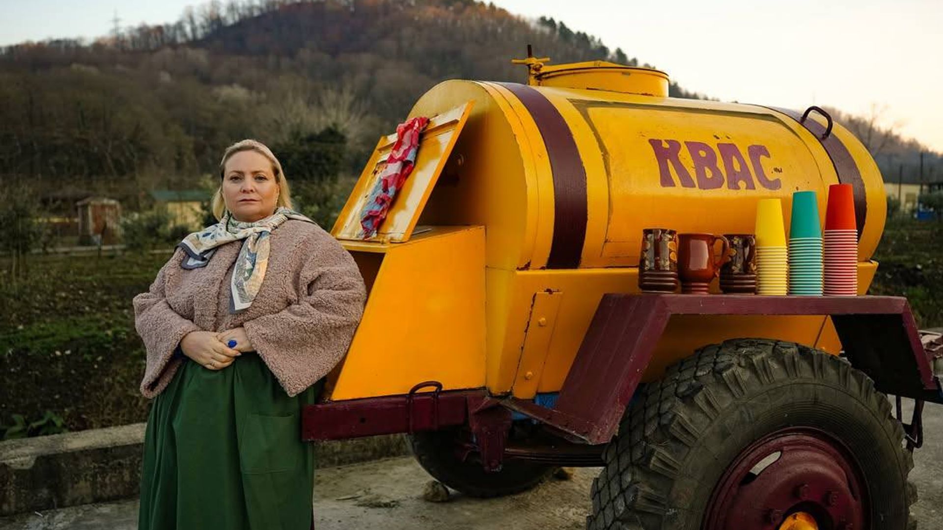 Светлана Пермякова в сельской пёстрой одежде стоит на фоне цистерны с квасом