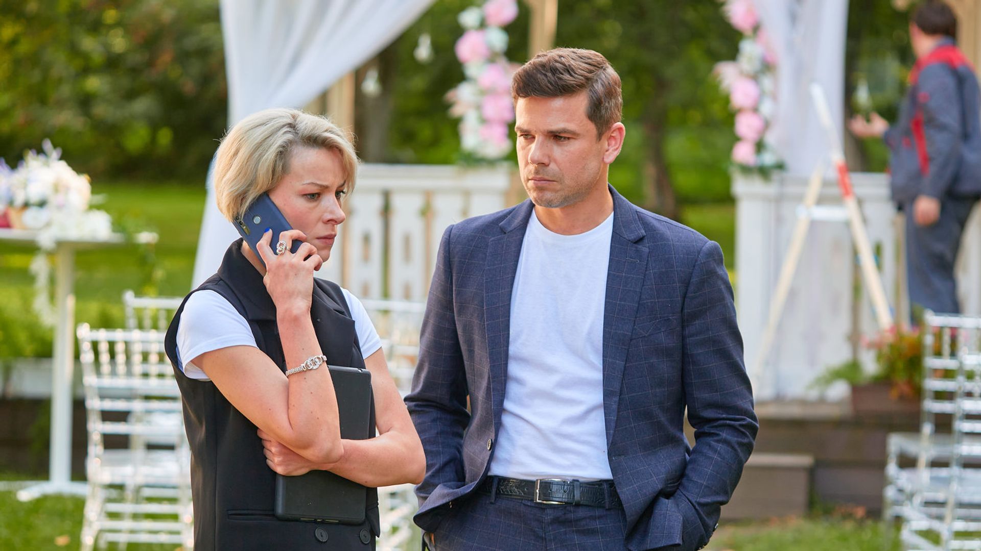 Женщина с короткими светлыми волосами говорит по телефону, рядом стоит мужчина в костюме на улице