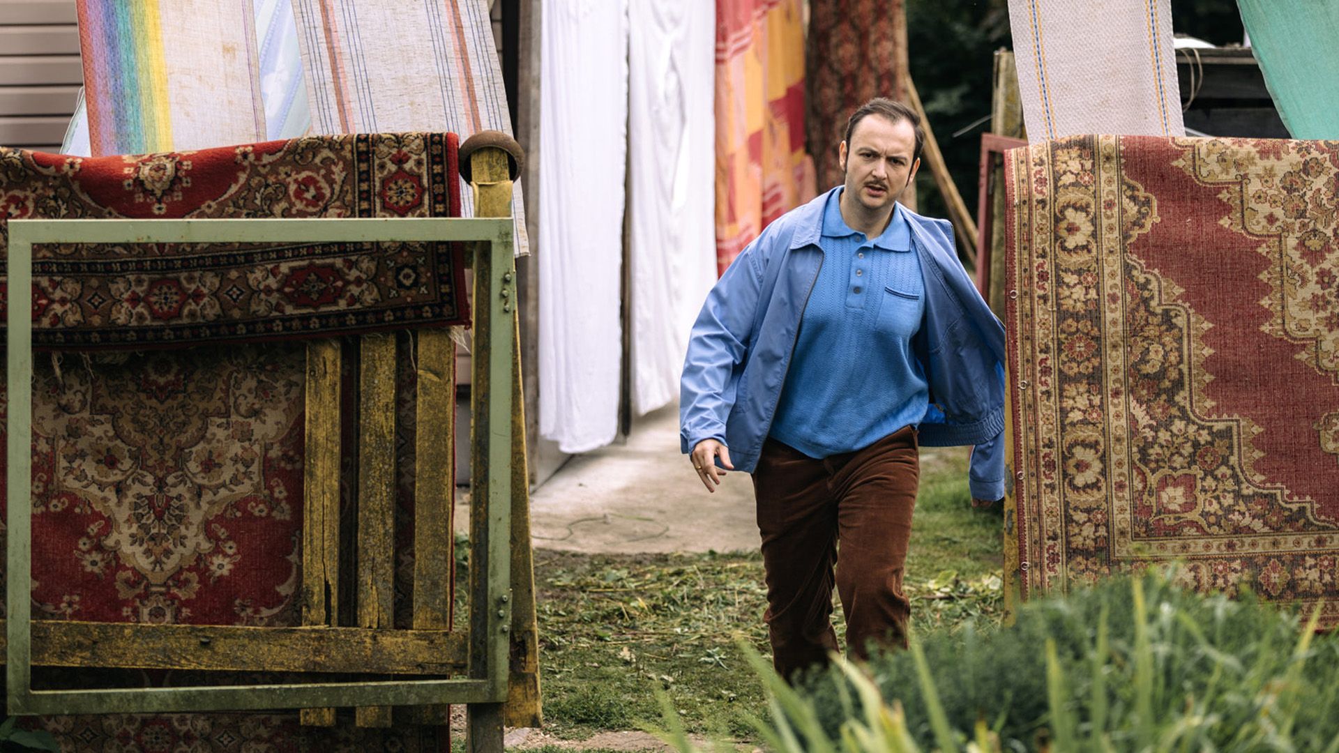 Алексей Золотовицкий в коричневых брюках и голубой куртке идёт по траве мимо висящих на заборе ковров