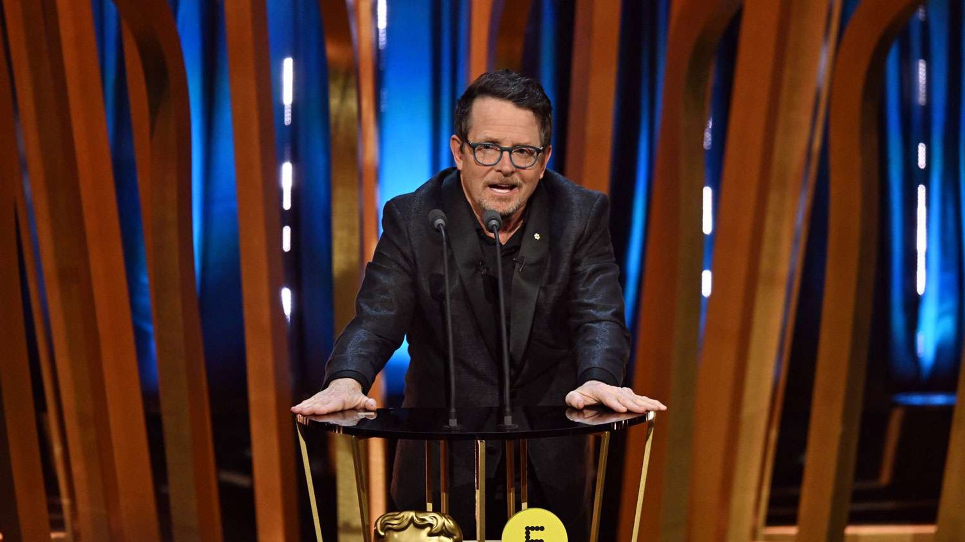 Майкл Джей Фокс в черном пиджаке говорит с трибуны