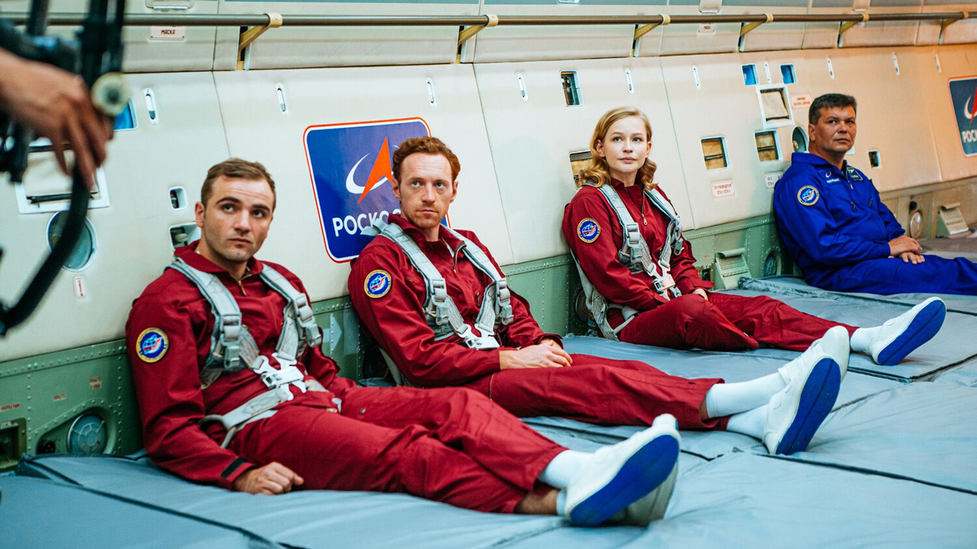 Максим Стоянов, мужчина и женщина, все в красных форменных комбинезонах, сидят на полу, прислонившись к стене с эмблемами "Роскосмоса"