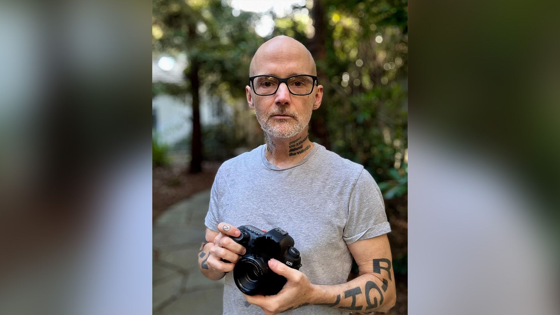 Моби в очках и серой футболке стоит в саду, держа в руках фотоаппарат