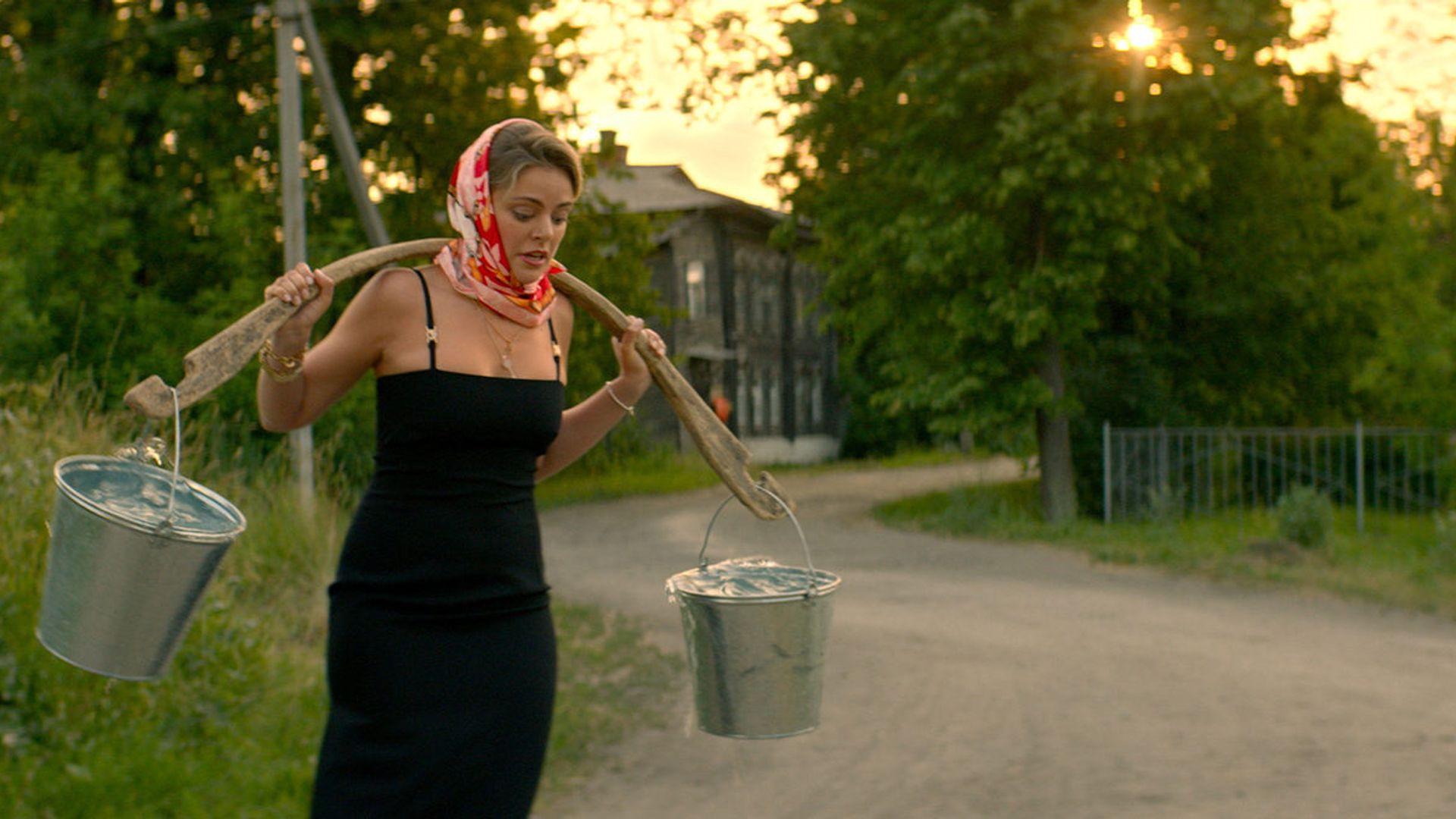 Маргарита Аброськина в платье и платке на голове несет коромысло с вёдрами