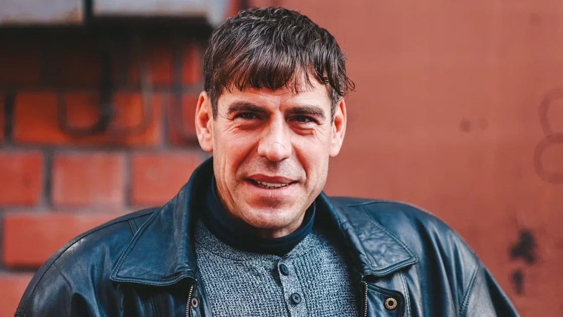 Дмитрий Дюжев в кожаной куртке на фоне красной стены