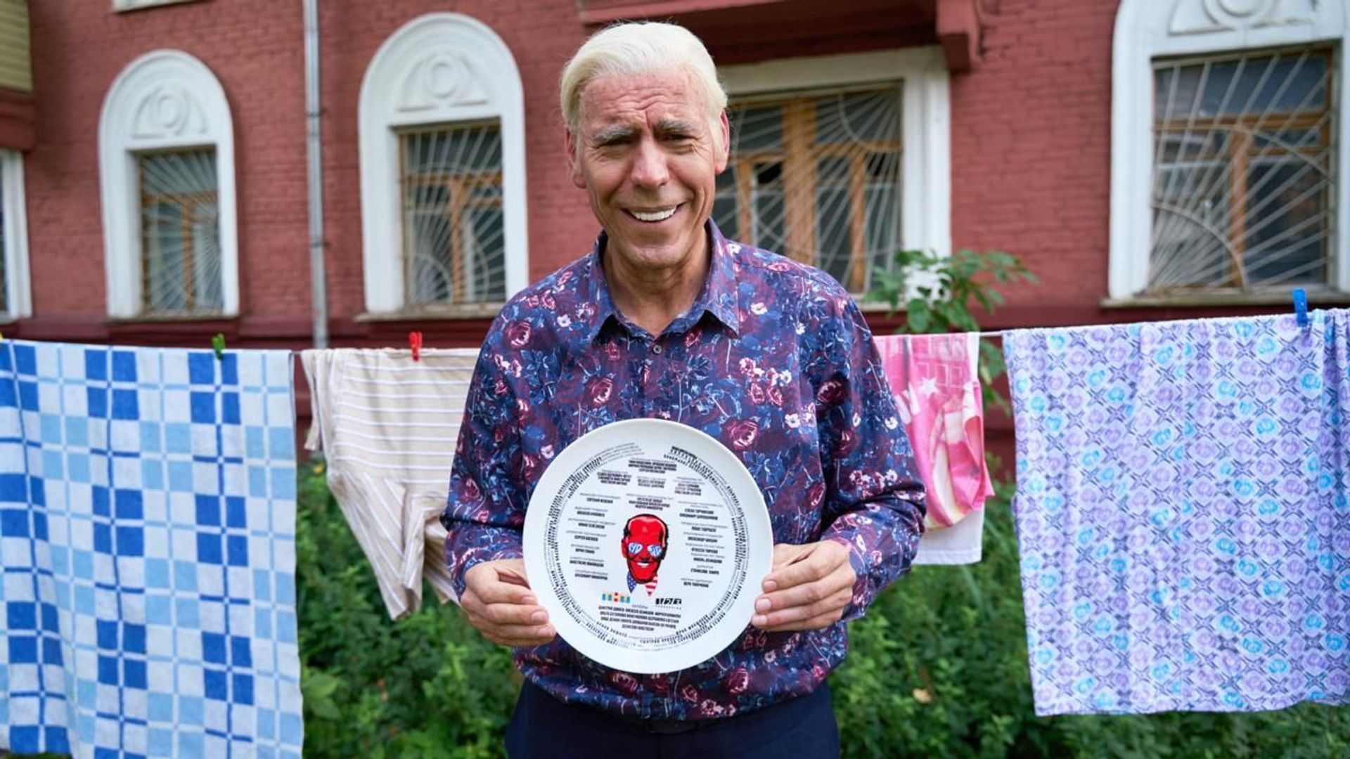 Дмитрий Дюжев загримированный под Байдена держит в руках тарелку с надписью Гудбай америка